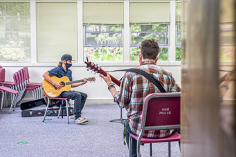 Two students playing guitar with masks and sitting in chairs on opposite sides of the room.