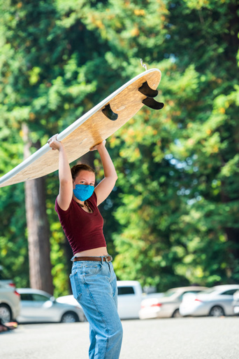 Student holding a surfboard over her head wearing a mask