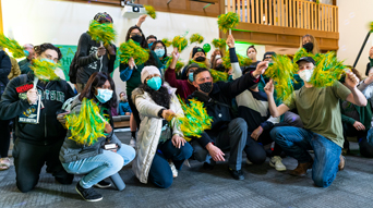 Students celebrating with green and gold pom poms in the SAC wearing masks