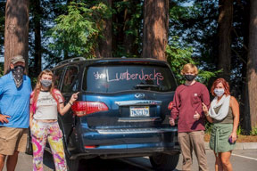 a family of four wearing masks posing with car and a lumberjack sign on the back window