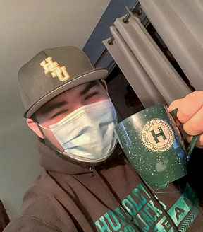 Student wearing a mask, a Green & Gold hat and sweatshirt, and holding an HSU mug.