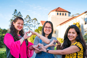 Thee students making hearts with their hands together in front of Founders Hall