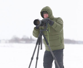 Mike with a big puffy green hooded jacket in front of a camera in snowy weather